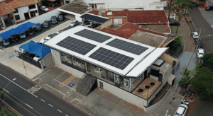 Vista aérea de um prédio comercial moderno com painéis solares no telhado, ao lado de um estacionamento com carros cobertos por tendas azuis, localizado em uma esquina urbana.