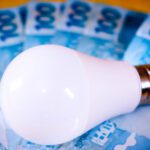 Lâmpada LED branca sobre várias cédulas de 100 reais dispostas em leque, simbolizando o aumento no custo da energia elétrica.
