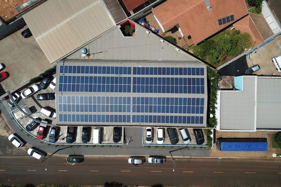 DCIM100MEDIADJI_0082.JPG Vista aérea de um prédio comercial com painéis solares instalados no telhado e estacionamento com carros ao redor.