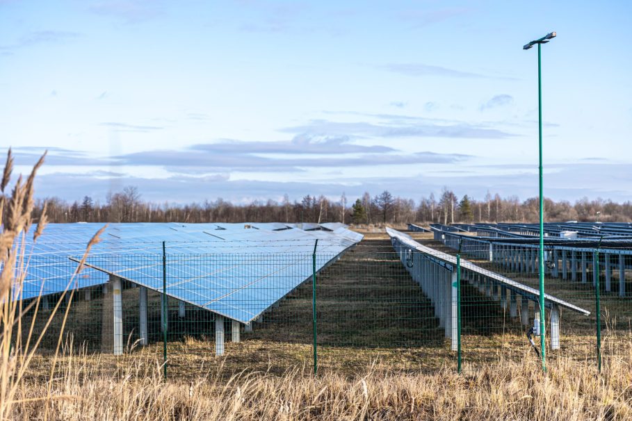Parque solar com fileiras de painéis fotovoltaicos instalados em um campo cercado, em dia claro com céu azul e nuvens.
