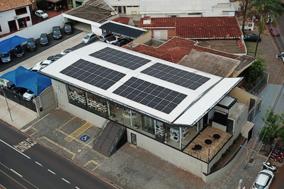 las-case-lojas-milano Vista aérea de um prédio comercial moderno com painéis solares no telhado, ao lado de um estacionamento com carros cobertos por tendas azuis, localizado em uma esquina urbana.