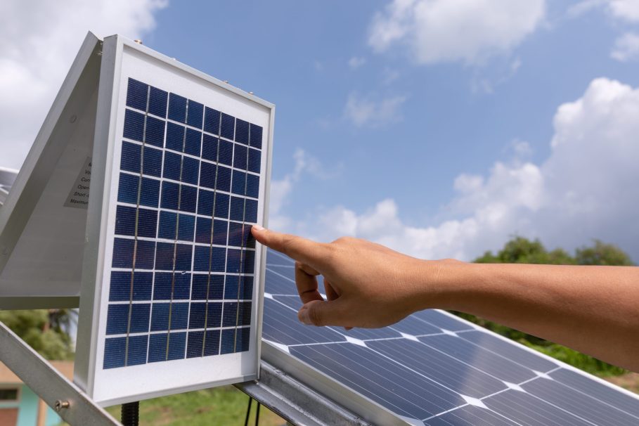 Engineer solar photovoltaic panels station checks Mão apontando para um painel solar fotovoltaico em um ambiente ao ar livre, com céu azul, nuvens leves e vegetação ao fundo.