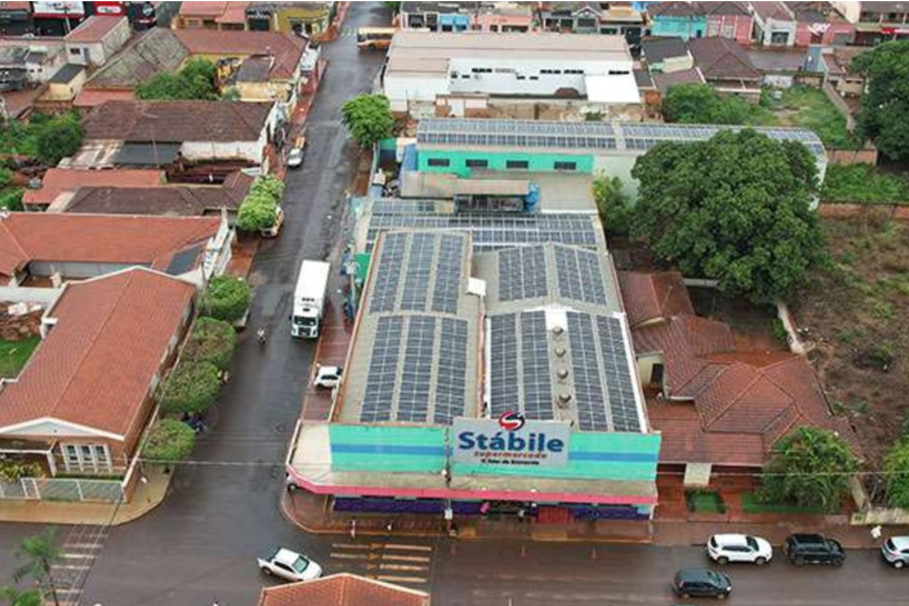 supermercados-miguelopolis Vista aérea de um supermercado chamado "Stábile" com placas solares no telhado, cercado por casas e árvores em uma área urbana.